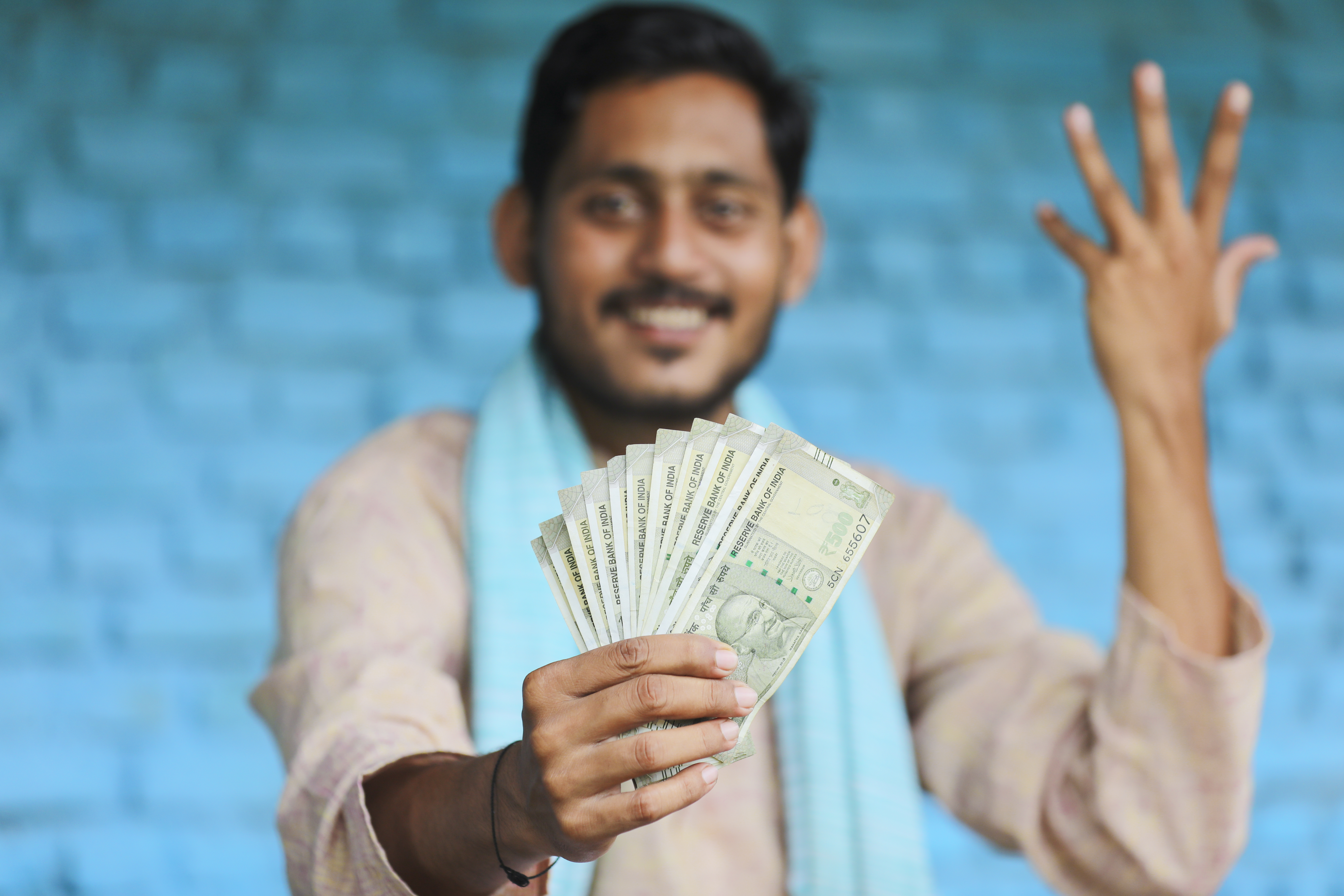 Man smiling, holding a fan of Indian rupee notes. Blurred blue brick wall in background, wearing light shirt and scarf. Mood: cheerful.