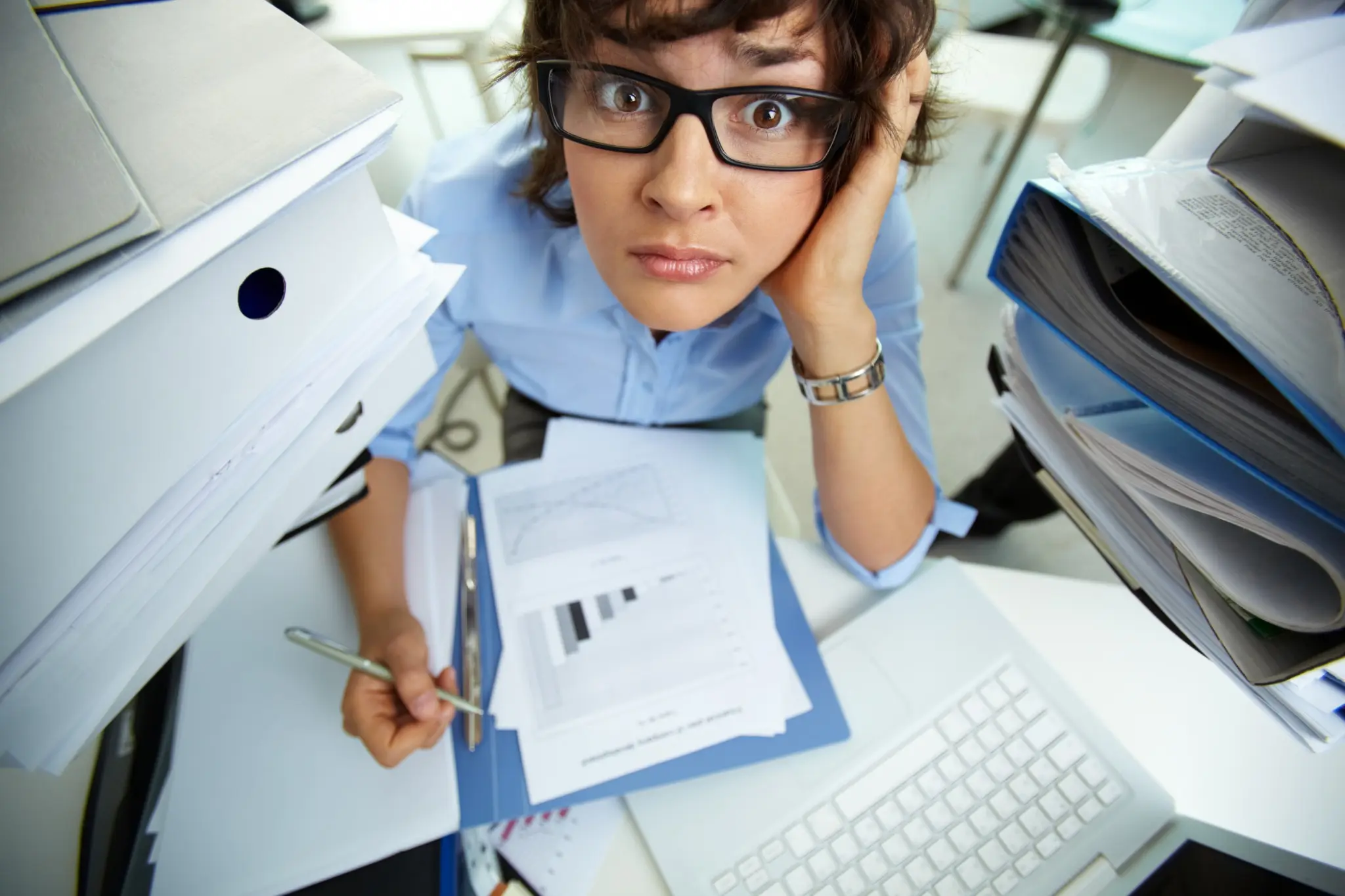 Woman in glasses looks stressed, surrounded by piles of documents and a computer. Papers with charts are on the desk in an office setting.